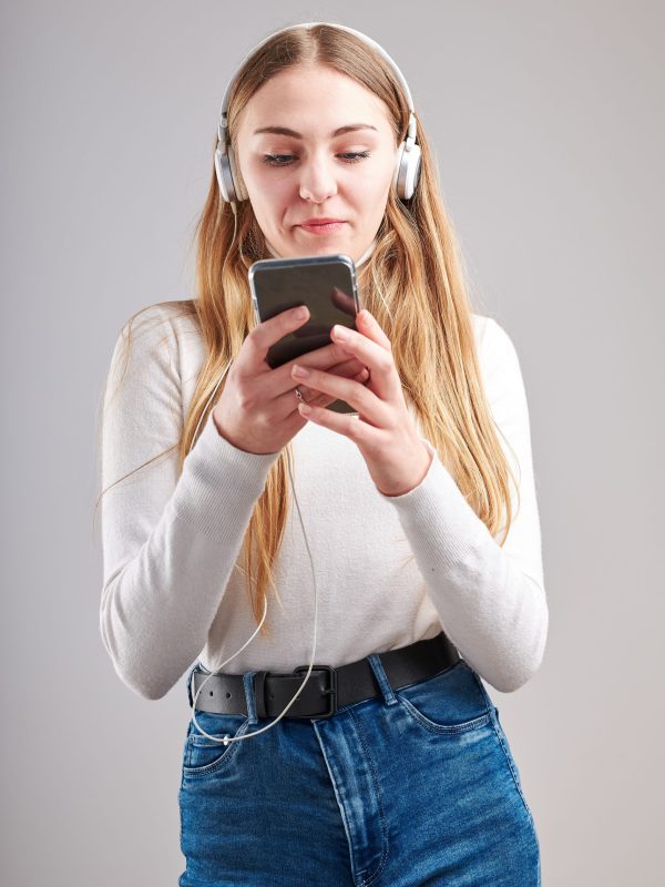 Young woman girl listening to music streaming content having fun watching video enjoying video chat talking with friends making gestures faces using smartphone earphones headphones standing over plain grey background